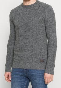 Grå stickad tröja med rund halsringning och ribbade muddar. Texturen är grov och har ett vävt mönster. Inkluderar en liten logotyp på fållen.