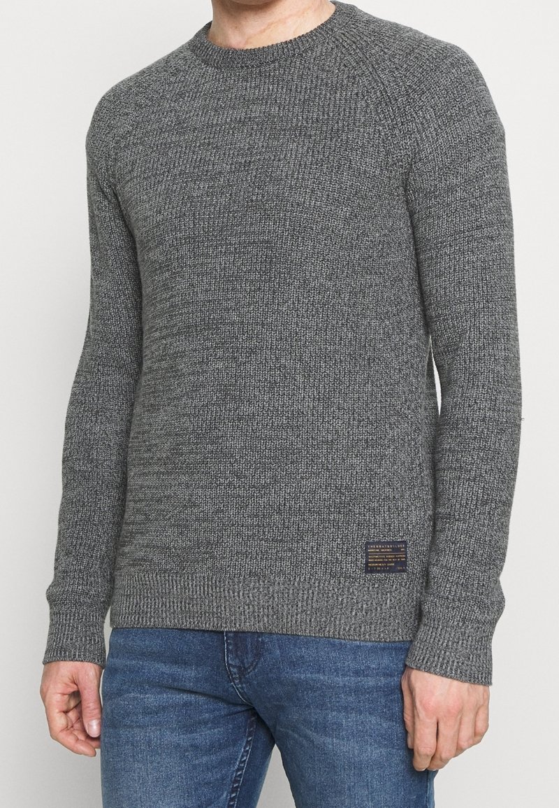 Grå stickad tröja med rund halsringning och ribbade muddar. Texturen är grov och har ett vävt mönster. Inkluderar en liten logotyp på fållen.