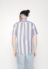 Krótki rękaw, koszula w paski w jasnym fioletowym i białym kolorze, z kołnierzykiem zapinanym na guziki i luźnym krojem. Miękka tkanina, widoczne szwy.