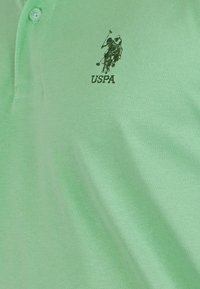 Zelena polo majica iz teksturiranega materiala, opremljena z dvema gumboma in izvezenim logotipom USPA v temnejši zeleni barvi na prsih.
