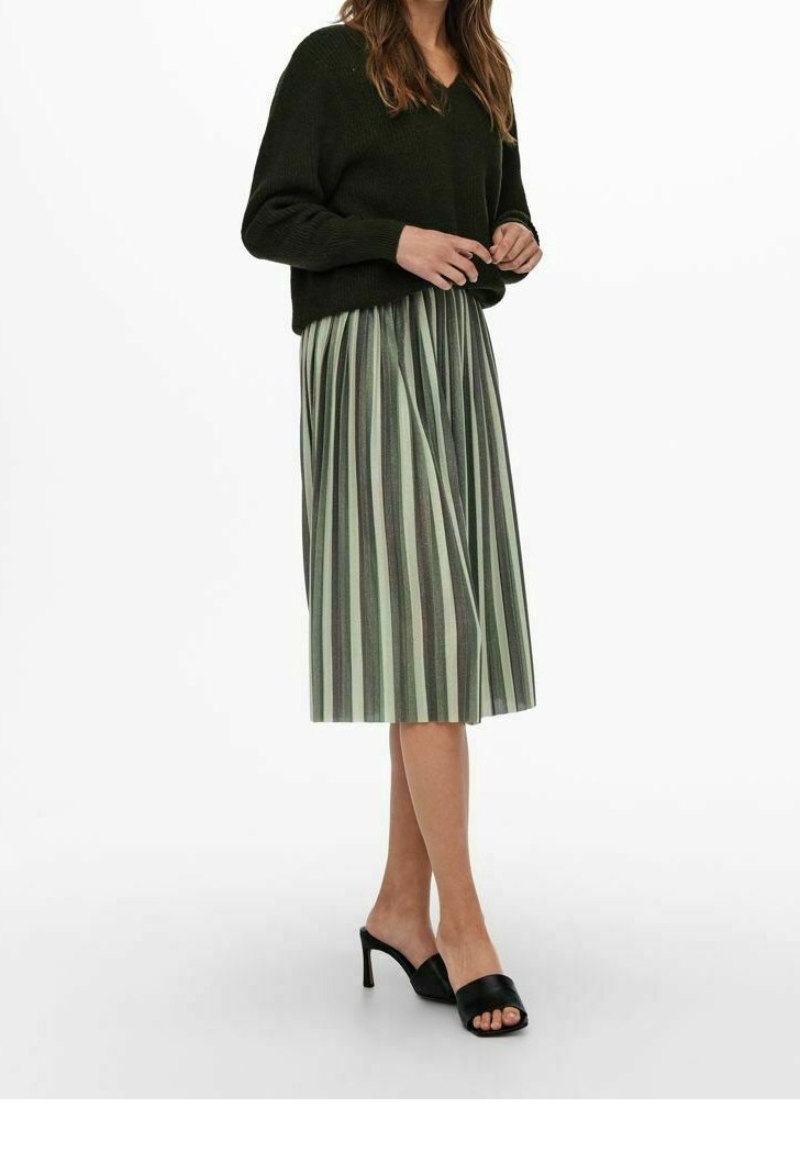 Femme portant un pull noir, une jupe midi plissée avec des rayures verticales vertes et grises, et des chaussures noires à talons hauts et bouts ouverts.