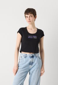 Crna skraćena majica s kratkim rukavima koja sadrži natpis "calvin klein od 1968" u bijeloj i ljubičastoj boji. Uparena s svijetloplavim trapericama.