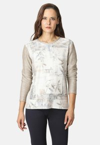 Mujer con cabello largo castaño, vistiendo una blusa de manga larga beige con estampado y pantalones negros, de pie frente a un fondo claro y liso.