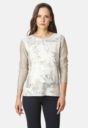 Mujer con cabello largo castaño, vistiendo una blusa de manga larga beige con estampado y pantalones negros, de pie frente a un fondo claro y liso.