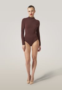 Kvinna som står klädd i en åtsittande långärmad brun bodysuit och beige högklackade skor mot en enfärgad ljus bakgrund.