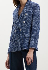 Femme portant un blazer croisé texturé bleu et blanc avec des boutons argentés, assorti à un jean bleu foncé, cropped aux épaules et à la taille.