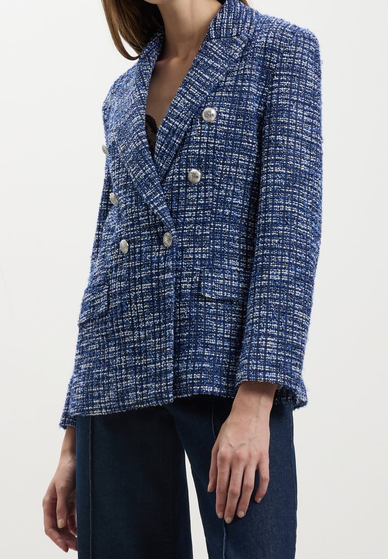 Femme portant un blazer croisé texturé bleu et blanc avec des boutons argentés, assorti à un jean bleu foncé, cropped aux épaules et à la taille.