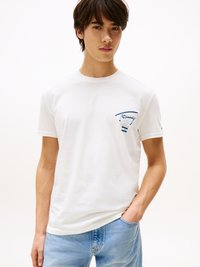 Vit bomull t-shirt med korta ärmar och rund hals, med en blå broderad logo på vänster bröst. Kombinerad med ljusblå jeans.