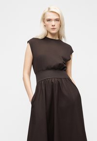 Ξανθιά γυναίκα που φοράει αμάνικο σκούρο καφέ φόρεμα με εφαρμοστή μέση και τσέπες, ποζάρει μπροστά σε λευκό φόντο.