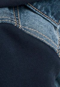 Denimová tkanina kombinovaná s námořnickou bavlnou. Obsahuje modré švy na denimu a ribbovanou texturu na bavlně. Detailní záběr na oblečení.
