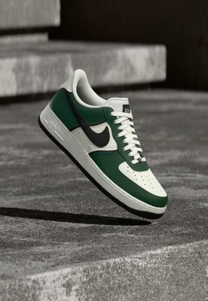 Grønne og hvide Nike Air Force 1 sneakers med sort swoosh, svævende over en betonoverflade med slørede trapper i baggrunden.