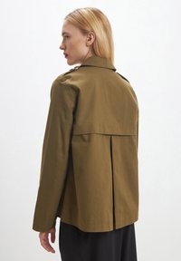 Donna con capelli biondi che indossa una giacca a maniche lunghe verde oliva, rivolta a sinistra su uno sfondo bianco semplice.