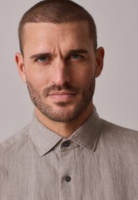 Hellgraues Leinenhemd mit Kragen, ausgestattet mit durchgehender Knopfleiste und strukturiertem Stoff. Das Modell hat eine kurz geschnittene Frisur und Gesichtshaare.