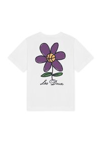Λευκό T-shirt με ένα μεγάλο μωβ λουλούδι στην πλάτη, πορτοκαλί μπάσκετ στο κέντρο του λουλουδιού, πράσινα φύλλα και το κείμενο "Les Deux" από κάτω.