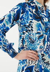 Blaues, floral gemustertes Hemd mit gepufften Ärmeln, geknöpften Manschetten und tailliertem Schnitt. Der Stoff wirkt glatt mit verschiedenen Blautönen und Designs.
