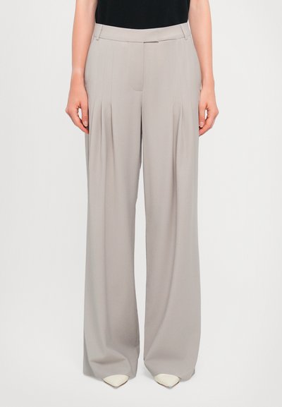 Pantalones de pierna ancha en gris claro con pliegues frontales prominentes, cintura ajustada y textura de tela suave. Combinados con tacones blancos puntiagudos.