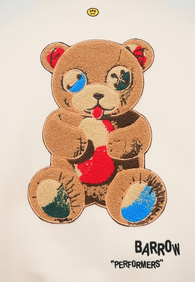 Bruin pluche teddybeer met meerkleurige vlekken en geborduurde details op een witte achtergrond, gelabeld "BARROW 'PERFORMERS'."