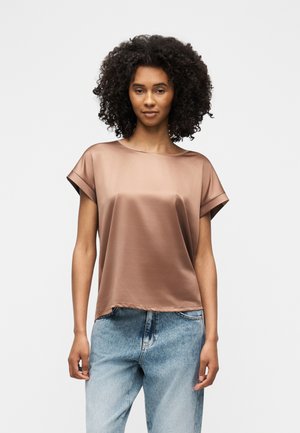 Γυναίκα με κατσαρά μαλλιά που φοράει κοντομάνικο λαμπερό καφέ τοπ και ανοιχτό μπλε τζιν, στέκεται μπροστά σε απλό ανοιχτό φόντο.