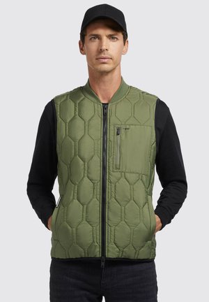 Grønne quiltede vest med hexagonalt mønster, frontlynlås og en lynlåslomme på brystet. Båret over en sort langærmet skjorte.
