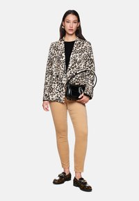 Leopardenmuster Blazer in Beige und Schwarz, kombiniert mit einem schwarzen Oberteil, hellbraunen Hosen und verzierten schwarzen Schuhen. Hält eine kleine schwarze Handtasche.