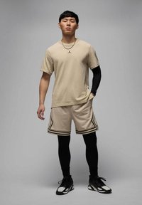 Camiseta atlética beige con logo, pantalones cortos beige con acentos negros y leggings negros. Modelo lleva zapatos deportivos negros y collares de plata.