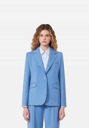 Giovane donna con capelli ricci che indossa un completo azzurro chiaro e una camicia a righe con bottoni, in piedi davanti a uno sfondo bianco semplice.