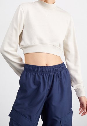 Personne portant un top court à manches longues crème et un pantalon cargo bleu marine à taille élastique avec poches, debout devant un fond uni.