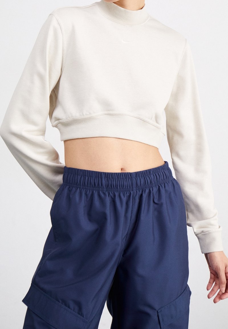 Personne portant un top court à manches longues crème et un pantalon cargo bleu marine à taille élastique avec poches, debout devant un fond uni.