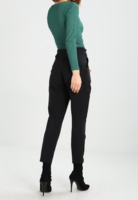 Donna che indossa un top verde a maniche lunghe, pantaloni neri su misura e stivaletti neri con tacco alto, in piedi con la schiena rivolta verso la fotocamera.