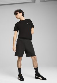 Camiseta atlética negra con logo, pantalones cortos negros a juego, calcetas negras y zapatillas negras con acentos dorados. Tela suave, corte relajado.