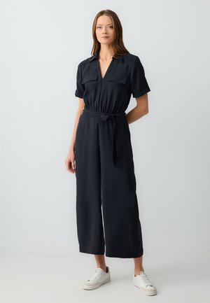 Combinaison marine à manches courtes, avec deux poches poitrine, fermeture éclair et ceinture à la taille. Jambes droites et tissu doux. Associée à des baskets blanches.