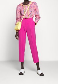 Rosa blus med blommönster och volangkrage, tillsammans med ljusrosa byxor. Accessoarer inkluderar en liten gul crossbody-väska och vita slides.