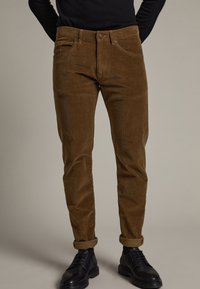 Corduroy trousers in rich brown, slim fit, featuring a classic waistband, five pockets, and rolled cuffs, paired with black shoes.