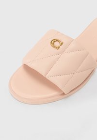 Pink slip-on sandal featuring a quilted strap, smooth sole, and a gold accent detail resembling a horseshoe. Made of faux leather.