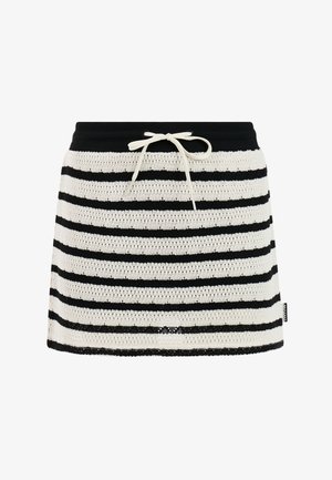 Mädchen mit blondem Haar steht mit den Händen in der Hüfte, trägt ein beigefarbenes T-Shirt, einen schwarz-weißen, gestreiften Rock mit Kordelzug und schwarze Flip-Flops.