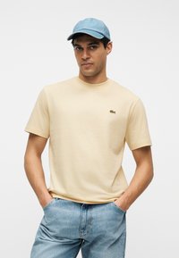 Άνδρας με μπεζ κοντομάνικη μπλούζα με πράσινο λογότυπο κροκόδειλου, μπλε τζιν τζιν και ανοιχτό μπλε καπέλο μπέιζμπολ, στέκεται με τα χέρια στις τσέπες.