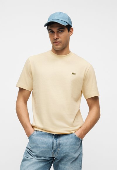 Homme portant un t-shirt beige à manches courtes avec un logo de crocodile vert, un jean en denim bleu et une casquette de baseball bleu clair, debout les mains dans les poches.