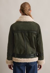 Groene suède jas met een crèmekleurige nepbont kraag en manchetten, een ceintuur in de taille en stikdetails langs de rug en mouwen.