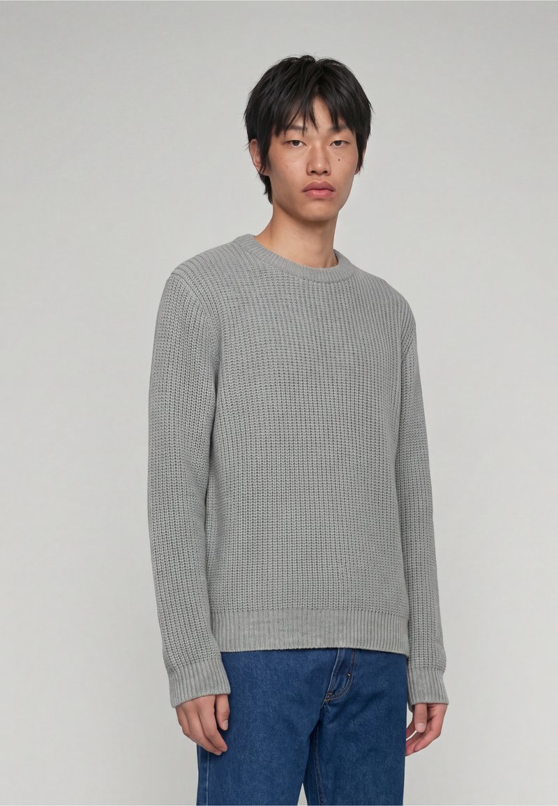 Jeune personne aux cheveux courts noirs, portant un pull en maille gris clair et un jean bleu, debout devant un fond uni.