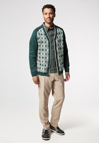 Zelený kardigan s bílým květinovým vzorem, zapnutý na knoflíky, nošený přes černou košili s potiskem, béžové kalhoty a černé tenisky.