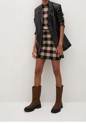 Bruine kniehoge laarzen met een gestructureerd oppervlak en een zwarte rubberen zool, gedragen met een zwart leren blazer over een beige en zwart geruit jurk.