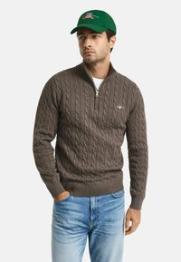 Brauner Strickpullover mit Reißverschluss und Kabelmuster, mit gerippten Bündchen und Saum. Trägt ein weißes Shirt mit blauen Jeans und einer grünen Mütze mit Stickerei.