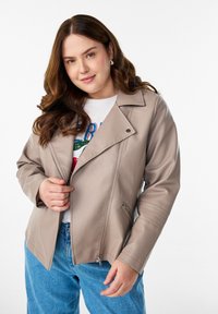 Beige Kunstlederjacke mit asymmetrischem Reißverschluss, breiten Revers und gesteppten Details. Kombiniert mit einem grafischen T-Shirt und blauen Jeans.