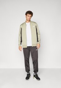 Lys grøn zip-up jakke med sorte sidepaneler, hvid T-shirt, mørkegrå joggers og sorte sneakers med grønne accenter.
