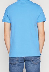 Světle modrá polo košile s krátkým rukávem a límečkem, vyrobená z měkké látky. Pohled zezadu ukazuje rovný lem a minimální detaily šití.