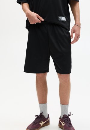 Schwarze Mesh-Sportshorts mit elastischem Bund, kombiniert mit einem locker sitzenden schwarzen Shirt und burgunderfarbenen Sneakers. Graue Socken werden getragen.