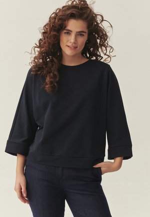 Mlada ženska z dolgimi kodrastimi lasmi, oblečena v široko temno modro majico in temne hlače, se smehlja z eno roko v žepu.