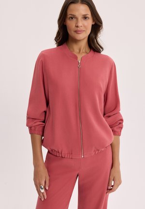 Kobieta ubrana w pasującą różowawą kurtkę z zamkiem błyskawicznym, z elastycznym obszyciem na dole i rękawach oraz dopasowane spodnie, stojąca na jasnym tle.