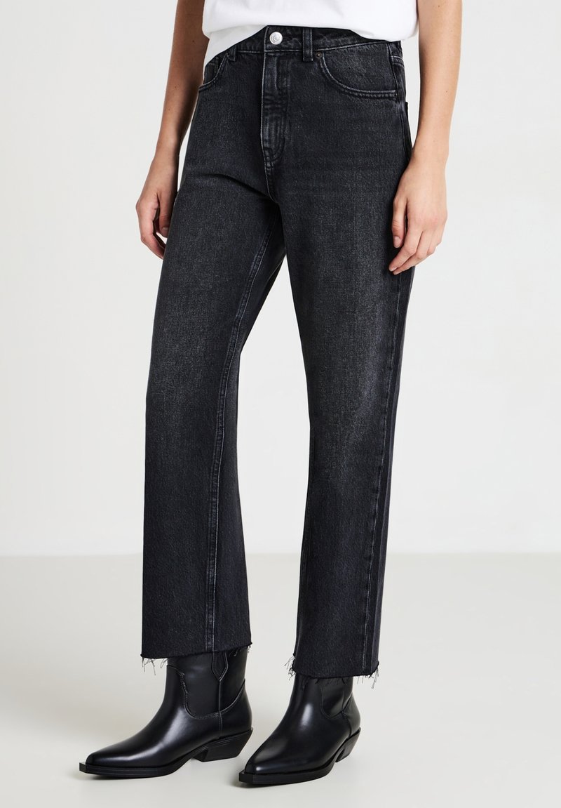 Jean en denim noir avec une coupe droite, présentant des ourlets effilochés. Porté avec des bottines noires à talons pointus. Design simple et épuré.