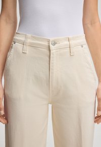Jeans beige chiari a vita alta con una texture liscia, dotati di chiusura con bottone in metallo e tasche frontali sottili.